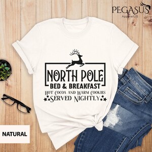 May include: White t-shirt with a black graphic design that says "North Pole Bed & Breakfast" with a reindeer illustration and the text "Hot cocoa and warm cookies served nightly".