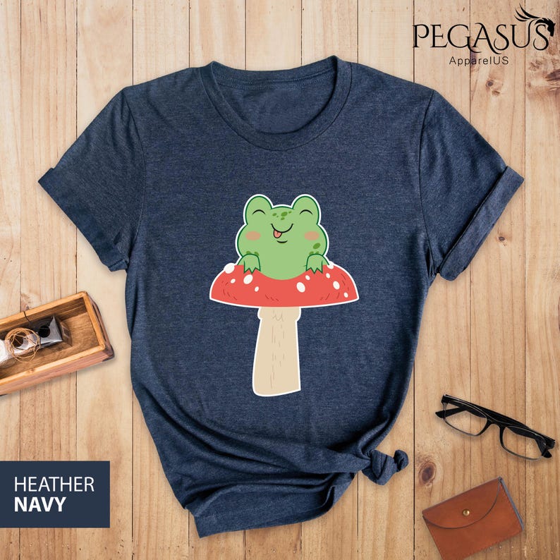May include: A dark blue t-shirt with a cartoon frog sitting on a red and white mushroom. The frog is green and has a big smile.