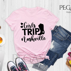 May include: Pink t-shirt with the text "Girls Trip Nashville" in black, featuring a heart and a cowboy boot graphic. The shirt is displayed with blue jeans, sunglasses, and a cup of tea.