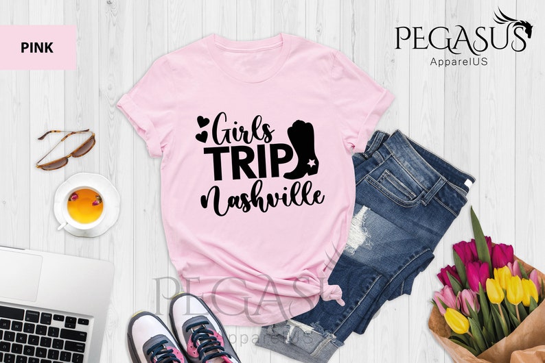 May include: Pink t-shirt with the text "Girls Trip Nashville" in black, featuring a heart and a cowboy boot graphic. The shirt is displayed with blue jeans, sunglasses, and a cup of tea.