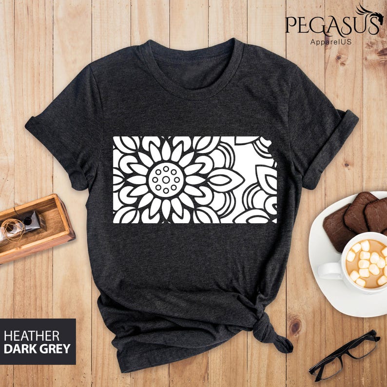 May include: A dark heather grey t-shirt with a white mandala design. The design is a large flower with intricate details. The t-shirt is laid flat on a wooden surface.