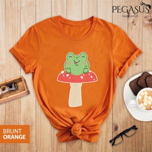 May include: Orange t-shirt with a cartoon frog sitting on a red and white mushroom. The text "BRUNT ORANGE" is at the bottom of the image.