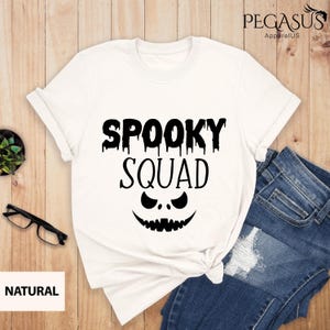 May include: White t-shirt with the text "SPOOKY SQUAD" in black, featuring a jack-o'-lantern face design. The shirt is folded, with a pair of blue jeans visible. A small plant and glasses are on the side.