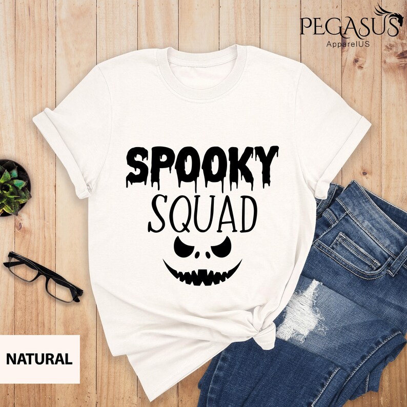 May include: White t-shirt with the text "SPOOKY SQUAD" in black, featuring a jack-o'-lantern face design. The shirt is folded, with a pair of blue jeans visible. A small plant and glasses are on the side.