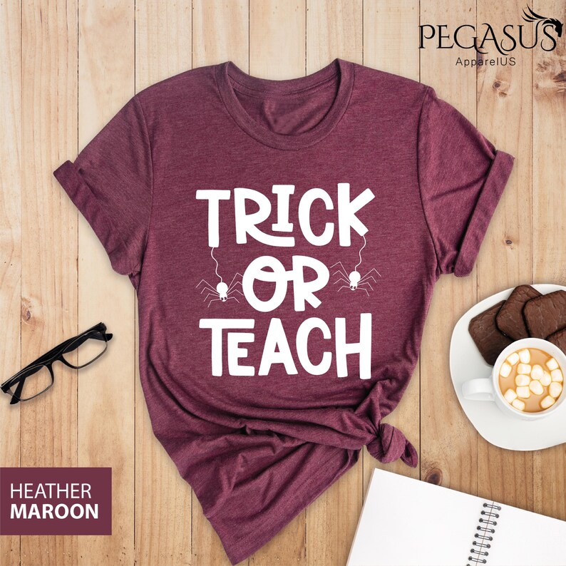 May include: Heather maroon t-shirt with the text "TRICK OR TEACH" in white, with spider graphics. The shirt is styled with rolled-up sleeves and a knotted hem. A pair of black glasses, a notepad, and a cup of cocoa are on a wooden surface.