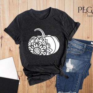 May include: A dark heather grey t-shirt featuring a white graphic of a pumpkin with a mandala design. The t-shirt is laid out on a wooden surface with a pair of jeans, a notebook, and a cup of coffee.