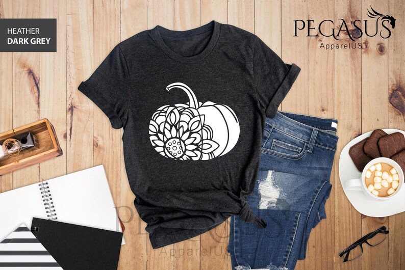 May include: A dark heather grey t-shirt featuring a white graphic of a pumpkin with a mandala design. The t-shirt is laid out on a wooden surface with a pair of jeans, a notebook, and a cup of coffee.
