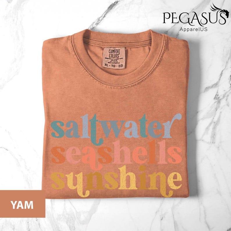 May include: A folded yam-colored t-shirt with the words "saltwater seashells sunshine" in colorful text. The shirt has a crew neck and a tag that reads "Comfort Colors". The Pegasus ApparelUS logo is in the upper right corner.