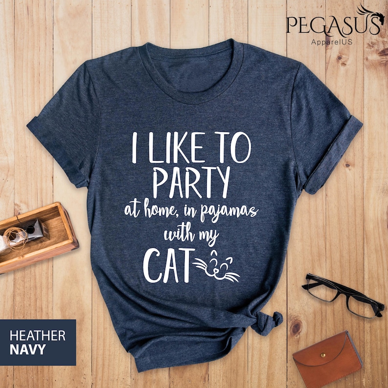 Op de afbeelding: Een heideblauw t-shirt met de tekst "Ik vind het leuk om te feesten thuis, in pyjama met mijn KAT" in wit. De tekst wordt vergezeld door een witte kattenkopgrafiek.