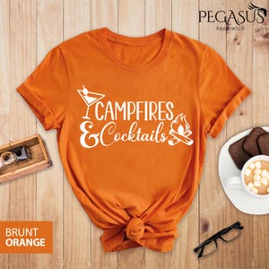 May include: Burnt orange t-shirt with the text "Campfires & Cocktails" in white. The design includes a cocktail glass and a campfire graphic. The shirt is knotted at the bottom and is on a wooden surface.