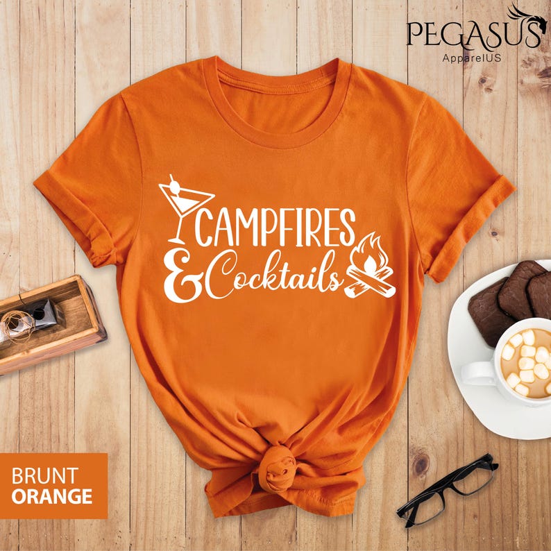 May include: Burnt orange t-shirt with the text "Campfires & Cocktails" in white. The design includes a cocktail glass and a campfire graphic. The shirt is knotted at the bottom and is on a wooden surface.