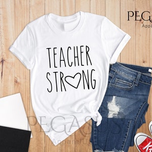 May include: White t-shirt with the text "TEACHER STRONG" and a heart in the middle of the word "STRONG". The shirt is laid out on a wooden surface with a pair of blue jeans and red sneakers.