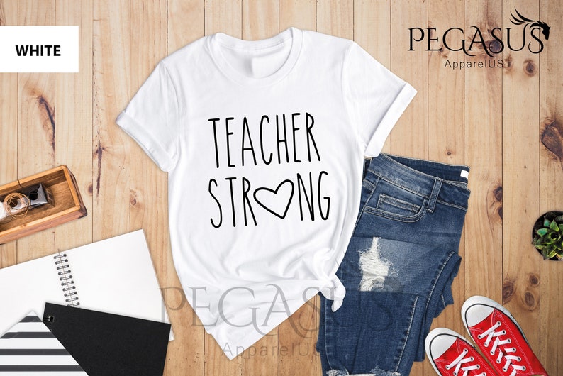 May include: White t-shirt with the text "TEACHER STRONG" and a heart in the middle of the word "STRONG". The shirt is laid out on a wooden surface with a pair of blue jeans and red sneakers.