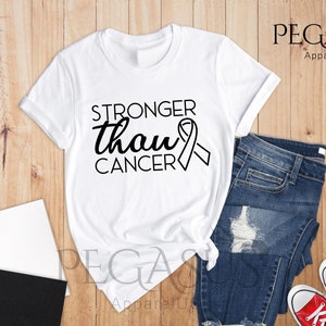 May include: White t-shirt with the text "Stronger than cancer" and a black ribbon graphic. The shirt is laid out on a wooden surface with a pair of blue jeans and red sneakers.