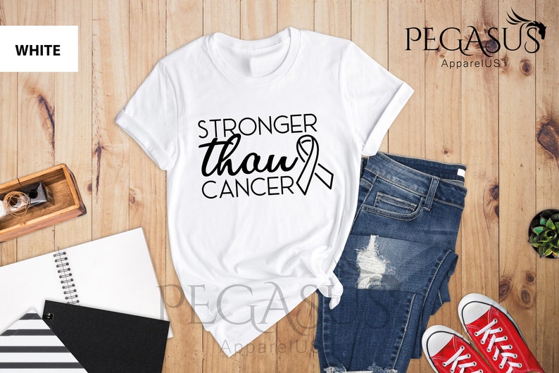 May include: White t-shirt with the text "Stronger than cancer" and a black ribbon graphic. The shirt is laid out on a wooden surface with a pair of blue jeans and red sneakers.