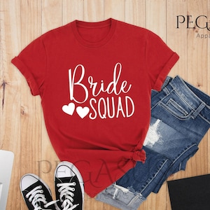 May include: Red t-shirt with the text "Bride Squad" in white with two white hearts. The shirt is laying on a wooden surface with a pair of black sneakers and a pair of blue denim shorts.