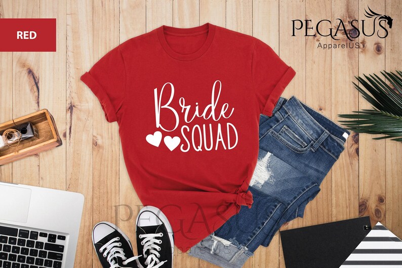 May include: Red t-shirt with the text "Bride Squad" in white with two white hearts. The shirt is laying on a wooden surface with a pair of black sneakers and a pair of blue denim shorts.