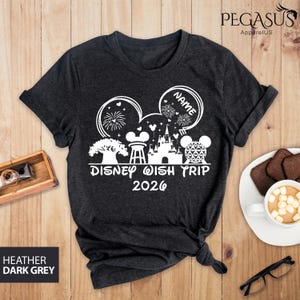 May include: Dark heather gray t-shirt with a white graphic design. The design includes Mickey Mouse ears, a castle, and the text "Disney Wish Trip 2026". The shirt also has a space to add a name. The shirt is made of a soft material.
