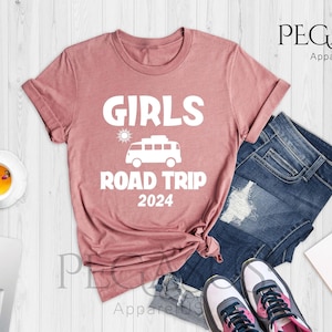 Puede incluir: Una camiseta de color malva con el texto "Girls Road Trip 2024" y un gráfico de una furgoneta con un sol encima.