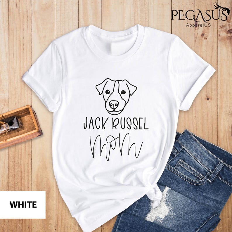 May include: White t-shirt with a black line drawing of a Jack Russell Terrier's face and the words "Jack Russel Mom" in black. The shirt is folded with a pair of blue jeans.