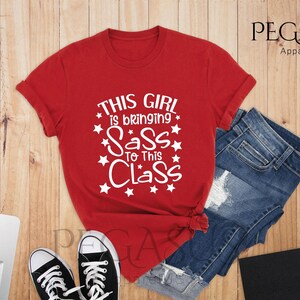 K&ouml;nnte beinhalten: Rotes T-Shirt mit wei&szlig;em Text, der "This girl is bringing sass to this class" lautet, umgeben von wei&szlig;en Sternen.