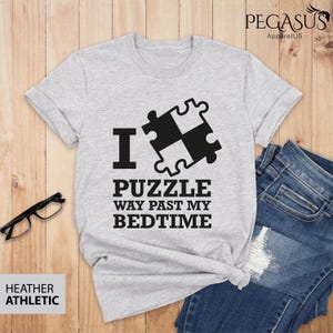 Puede incluir: Una camiseta gris jaspeada con un gr&aacute;fico negro de una pieza de rompecabezas y el texto "I &hearts; Puzzle Way Past My Bedtime".