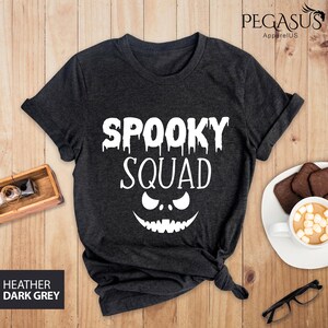 May include: Dark heather gray t-shirt with the words "SPOOKY SQUAD" in white, featuring a jack-o'-lantern face design. The shirt is a crew neck style. The text "HEATHER DARK GREY" is in a box in the lower left corner.