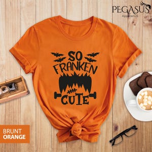 Pu&ograve; includere: T-shirt arancione bruciato con una grafica nera che recita "So Franken Cute" con pipistrelli e un design di mostro di Frankenstein. La maglietta &egrave; annodata in basso. Logo Pegasus ApparelUS in alto a destra.