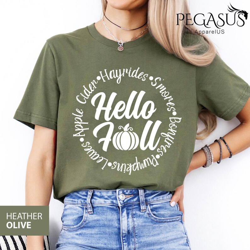 May include: Heather olive green t-shirt with the words "Hello Fall" in white, surrounded by a circle of fall-themed words like "Cider" and "Pumpkins". The shirt has short sleeves and is made of a soft material.