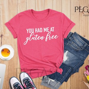 K&ouml;nnte beinhalten: Ein pinkfarbenes T-Shirt mit dem Text "You had me at gluten free" in wei&szlig;en Buchstaben. Das Shirt liegt auf einer Holzoberfl&auml;che mit einer Jeans, einer Tasse Tee, einem Laptop und einem Blumenstrau&szlig;.