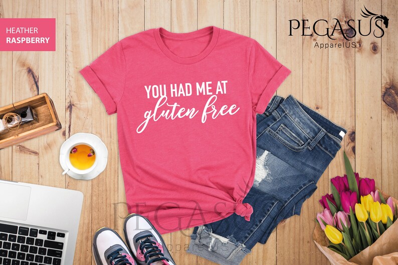 K&ouml;nnte beinhalten: Ein pinkfarbenes T-Shirt mit dem Text "You had me at gluten free" in wei&szlig;en Buchstaben. Das Shirt liegt auf einer Holzoberfl&auml;che mit einer Jeans, einer Tasse Tee, einem Laptop und einem Blumenstrau&szlig;.