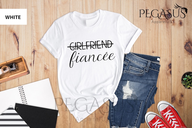 May include: White t-shirt with the text "GIRLFRIEND" crossed out and "fianc&eacute;e" written below it. The shirt is on a wooden surface with a pair of jeans and red sneakers.