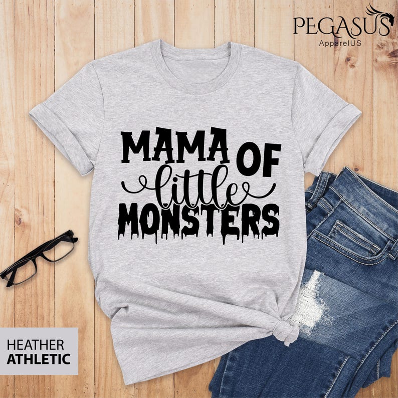 May include: A light gray heather athletic t-shirt with the text "MAMA OF little MONSTERS" in black. The shirt is styled with a knot at the waist and is paired with blue jeans. The shirt also has the text "HEATHER ATHLETIC".