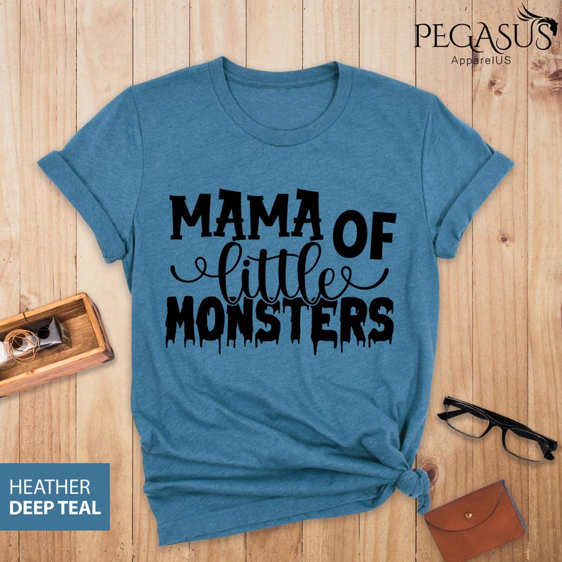 May include: A deep teal t-shirt with the phrase "MAMA OF little MONSTERS" in black script. The shirt has rolled-up sleeves and a knotted hem. The words "Heather Deep Teal" are in a blue box in the lower left corner.