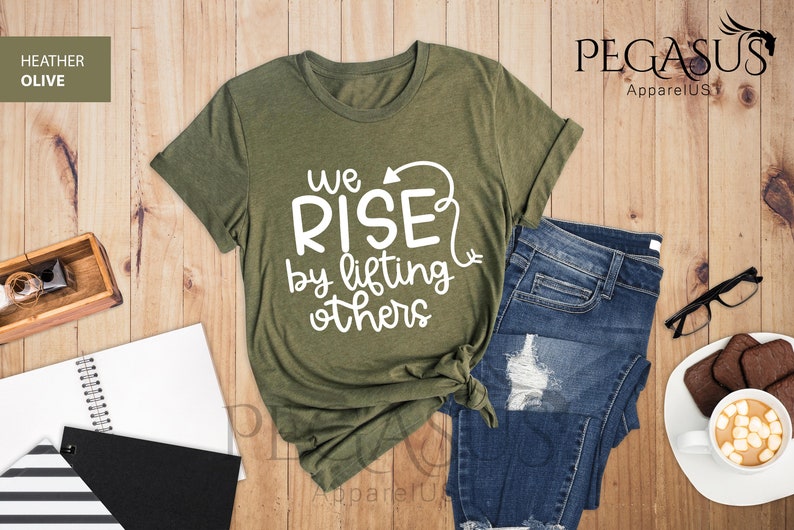 Pu&ograve; includere: Una maglietta verde in cotone heather oliva con il testo "We Rise by lifting others" in lettere bianche. La maglietta &egrave; piegata e mostrata su una superficie di legno con un paio di jeans, un quaderno e una tazza di caff&egrave;.