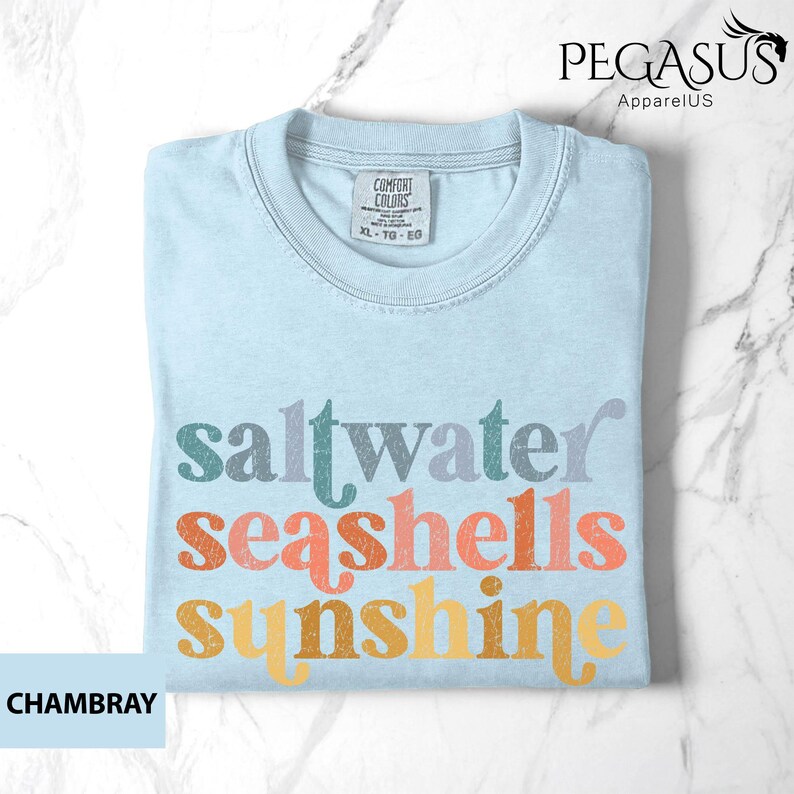 May include: Light blue t-shirt with the words "saltwater seashells sunshine" in colorful, retro-style lettering. The shirt has a crew neck and a tag that reads "Comfort Colors". The shirt is made of a soft material.