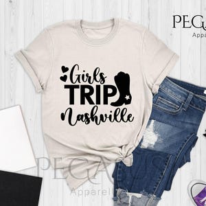 May include: A natural-colored t-shirt with the text "Girls Trip Nashville" in black, along with a black cowboy boot graphic. The shirt is styled with blue denim shorts and purple sneakers.