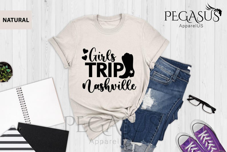 May include: A natural-colored t-shirt with the text "Girls Trip Nashville" in black, along with a black cowboy boot graphic. The shirt is styled with blue denim shorts and purple sneakers.