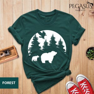 Puede incluir: Una camiseta verde oscuro con un gr&aacute;fico blanco de un oso, un oso m&aacute;s peque&ntilde;o y un p&aacute;jaro volando sobre un c&iacute;rculo de &aacute;rboles. El texto "FOREST" est&aacute; en la esquina inferior izquierda y "PEGASUS ApparelUS" est&aacute; en la esquina superior derecha.
