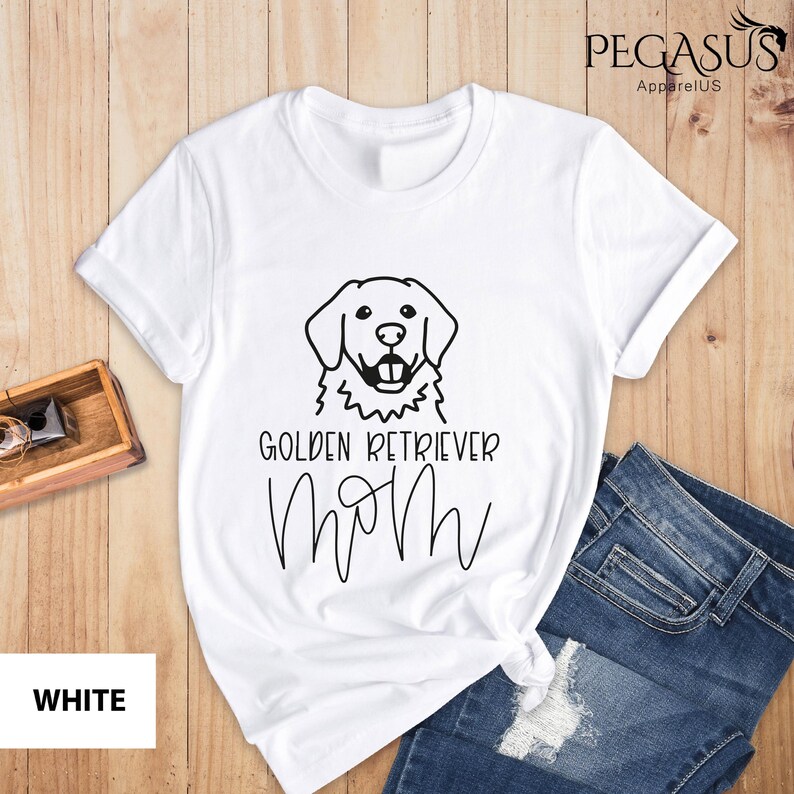 Op de afbeelding: Wit T-shirt met een zwarte omtrek van een golden retriever hond en de tekst "Golden Retriever Mom" in zwart.