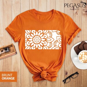 May include: A burnt orange t-shirt with a white mandala design. The design is a stylized sunflower with intricate details. The t-shirt is made of a soft, comfortable material.