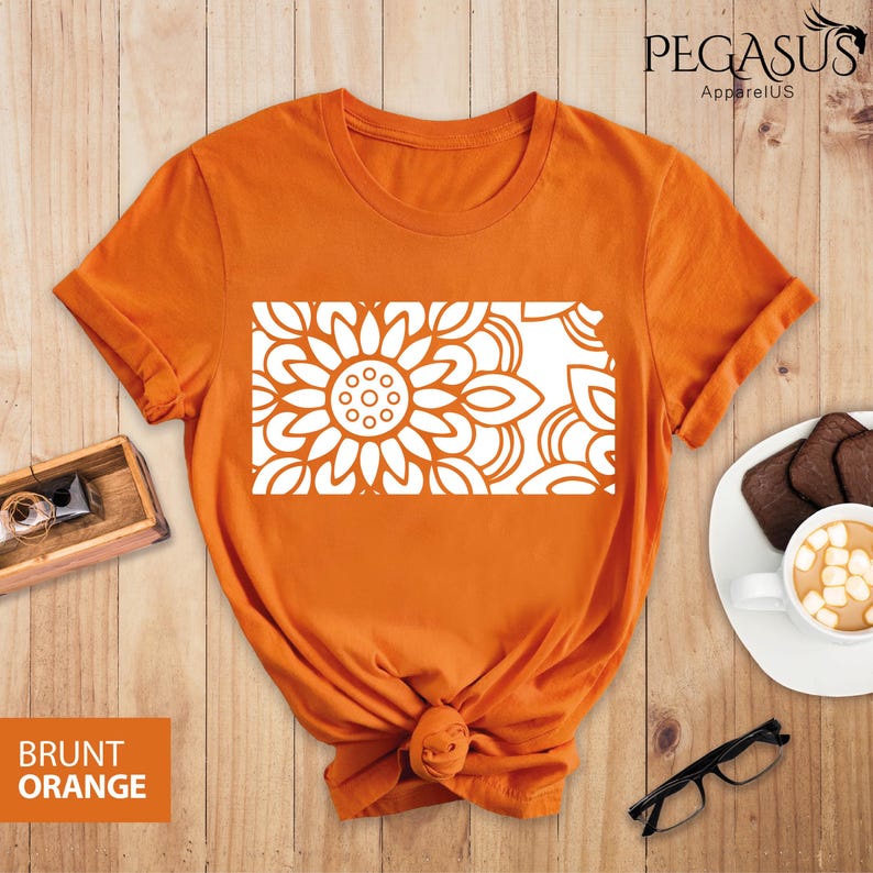May include: A burnt orange t-shirt with a white mandala design. The design is a stylized sunflower with intricate details. The t-shirt is made of a soft, comfortable material.
