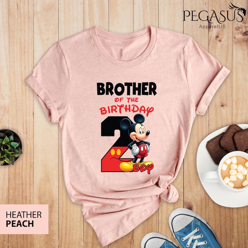 May include: Heather peach t-shirt with the text "BROTHER OF THE BIRTHDAY" and a graphic of Mickey Mouse with the number 2. The shirt is a crew neck and has short sleeves. The shirt is laying on a wooden surface.