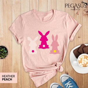 May include: Heather peach-colored t-shirt featuring three bunny silhouettes in pink and white. The bunnies have patterned designs and a yellow or white tail. The shirt is displayed on a wooden surface with a pair of blue sneakers and a cup of hot chocolate.