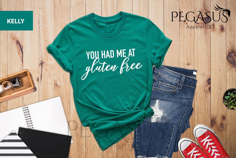 K&ouml;nnte beinhalten: Ein gr&uuml;nes T-Shirt mit dem Text "You had me at gluten free" in wei&szlig;er, kursiv geschriebener Schrift. Das Shirt liegt auf einer Holzoberfl&auml;che mit einer Jeans und roten Turnschuhen.