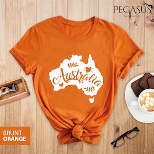 Op de afbeelding: Een gebrand oranje T-shirt met een witte omtrek van het Australische continent en het woord "Australia" in een gestileerd lettertype. Het ontwerp bevat ook kleine hartjes en bladeren.