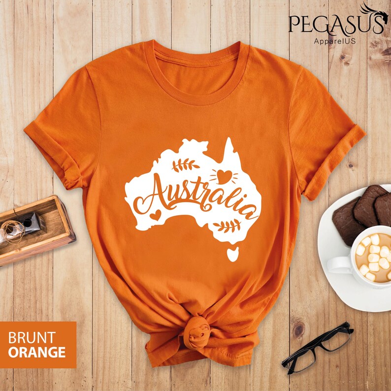 Op de afbeelding: Een gebrand oranje T-shirt met een witte omtrek van het Australische continent en het woord "Australia" in een gestileerd lettertype. Het ontwerp bevat ook kleine hartjes en bladeren.