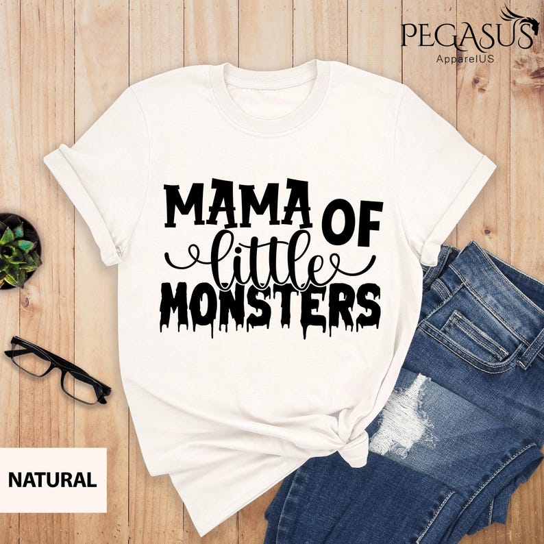 May include: A white t-shirt with the text "MAMA OF little MONSTERS" in black. The shirt is styled with rolled-up sleeves and a knot at the bottom. A pair of blue jeans is visible on the right side of the image.