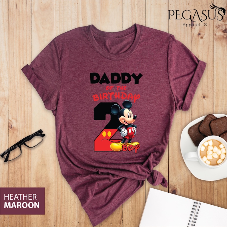 May include: Heather maroon t-shirt with the text "DADDY of the BIRTHDAY 2 BOY" and a Mickey Mouse graphic. The shirt has rolled-up sleeves and a knotted bottom. The shirt is on a wooden surface.