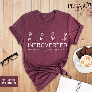 May include: Heather maroon t-shirt with white text that reads "Introverted but willing to discuss plants". The shirt features a line of four simple white flower illustrations.